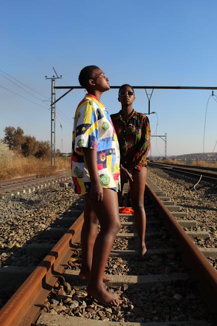 Two women standing confidently on Soweto railway tracks, embracing vibrant fashion under a clear sky.