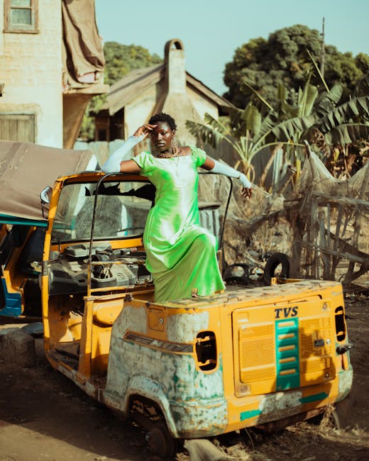 A fashionable woman in a vibrant green dress poses on a rustic vehicle in a rural setting.
