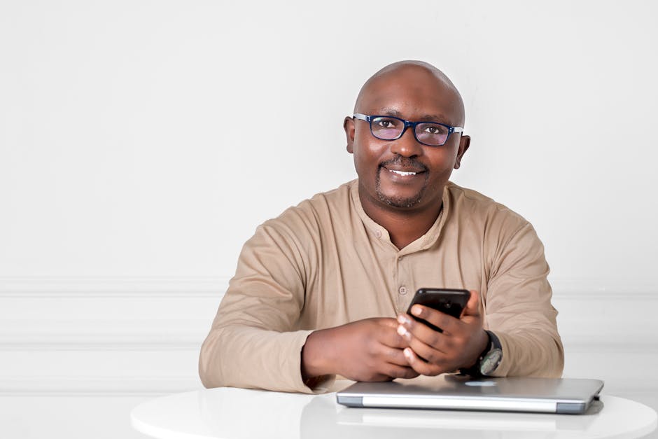 Smiling man in glasses using smartphone at table with laptop on a white background.