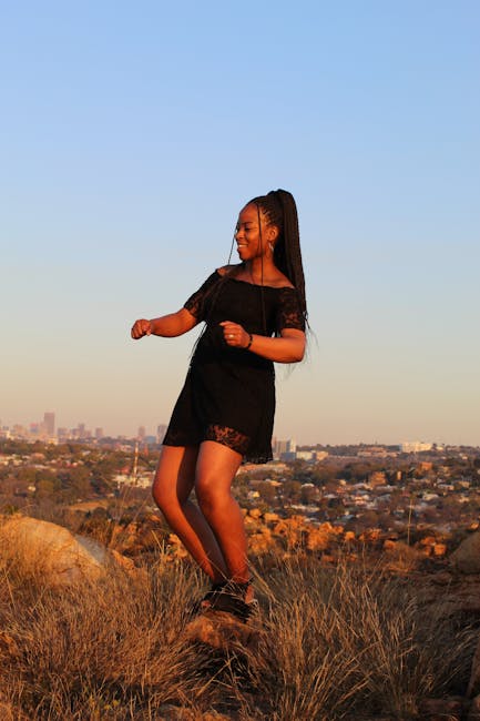 Black woman in a black dress dancing atop hills with a cityscape view at sunset.
