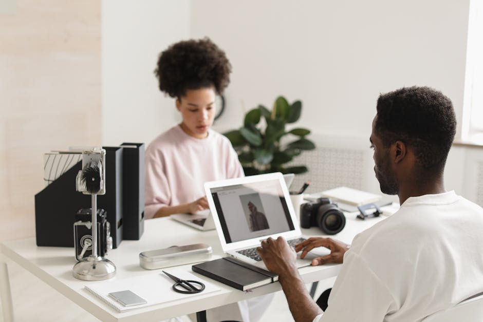 Two creative professionals collaborating in a bright, modern office setting with laptops and camera equipment.