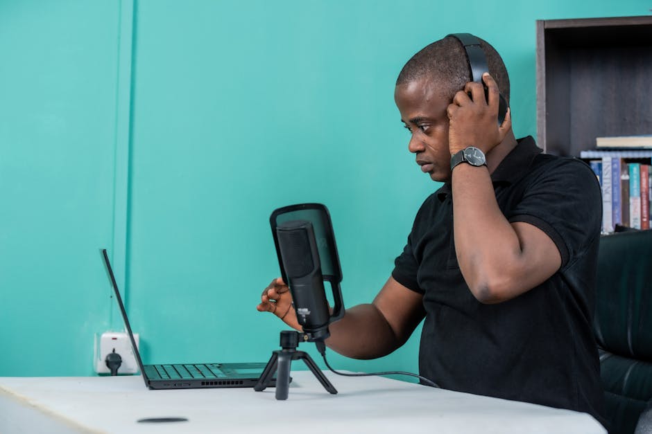Man in black shirt recording a podcast using laptop, microphone, and headphones.