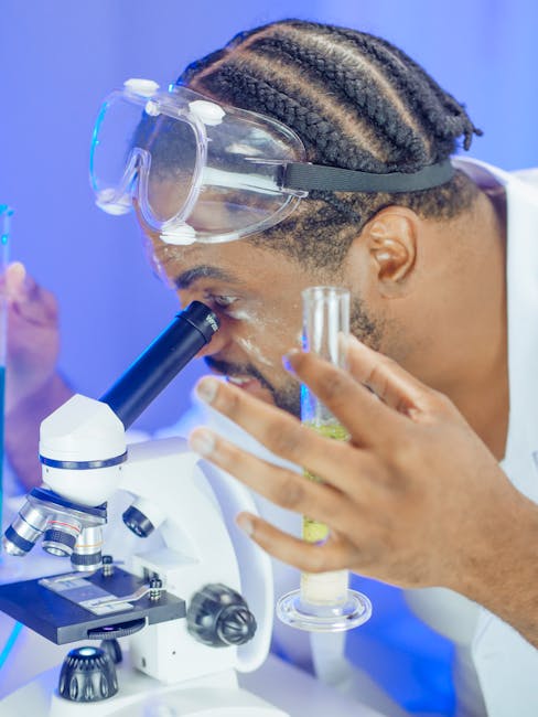 A scientist in a lab coat examining a sample under a microscope with protective goggles.