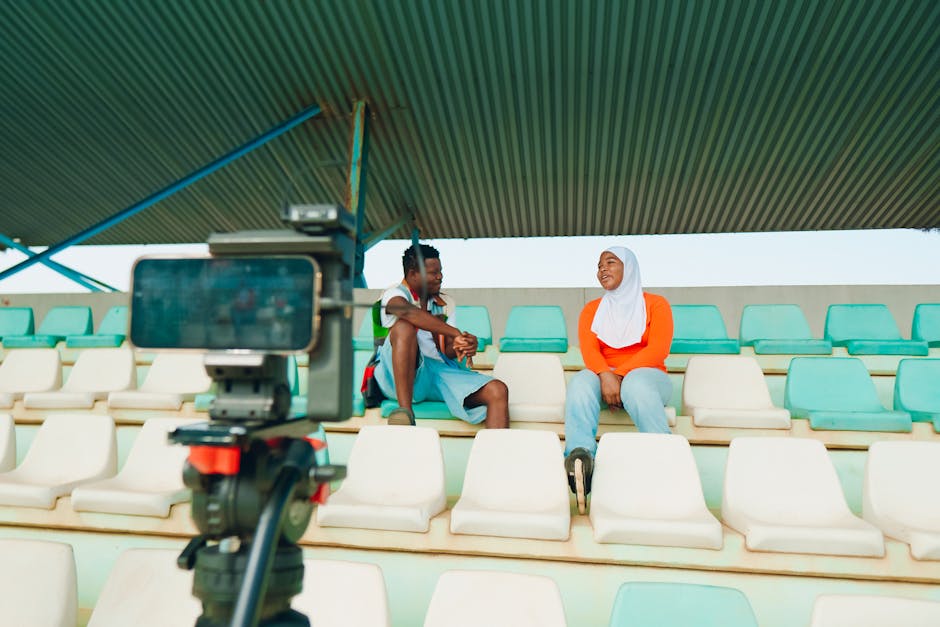 Two people conduct an interview in stadium seats with a video camera setup.