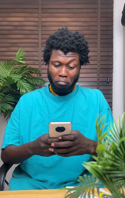 African man with thoughtful expression using smartphone indoors surrounded by plants.