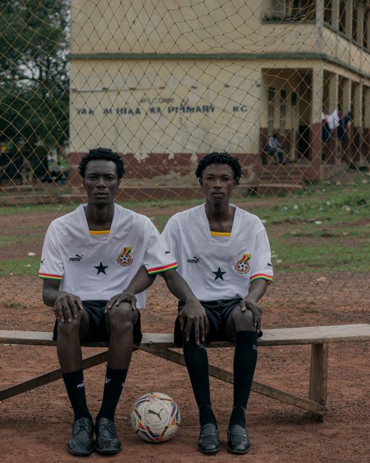 Two Ghanaian soccer players sitting on a bench with a ball on a dirt field.