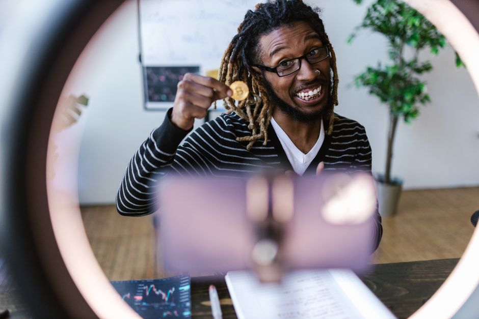 African American man smiling while holding a Bitcoin in front of a smartphone for a video recording.