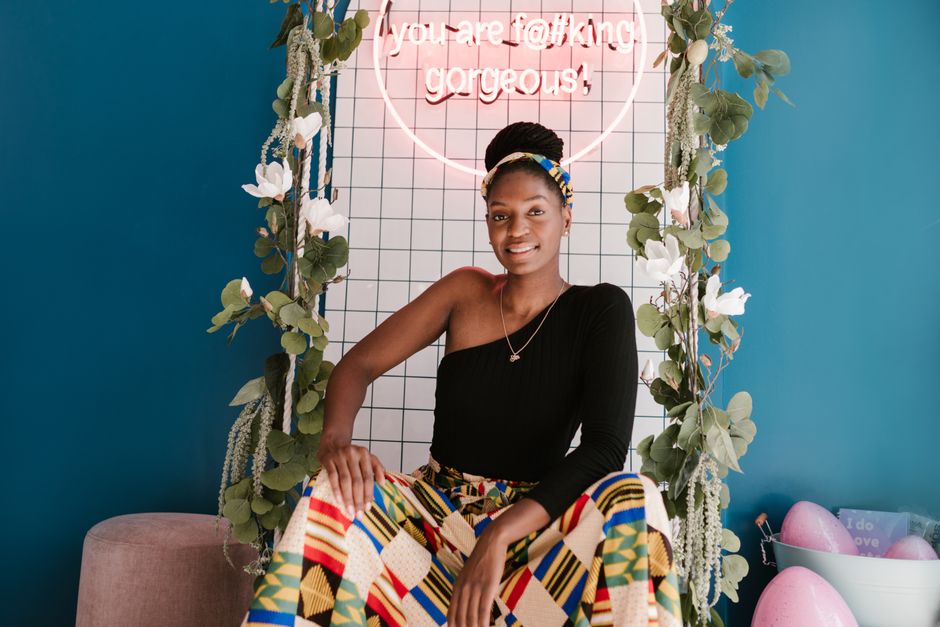 A smiling woman in colorful attire poses elegantly in a stylish indoor setting with neon lighting.